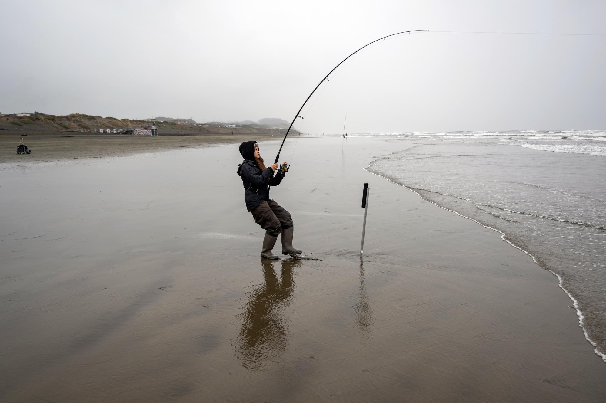 A woman on the beach fishes with a fishing pole on a foggy day.