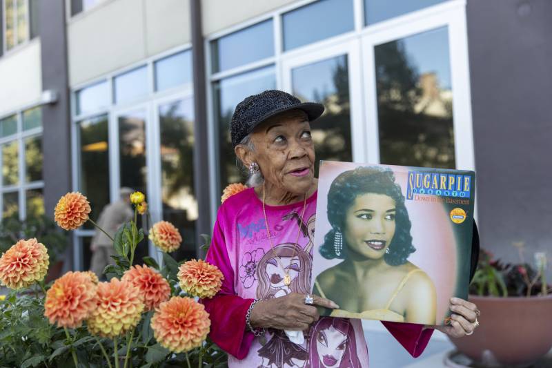 An 87-year-old woman with a pink top and sequined hat poses in front of flowers.