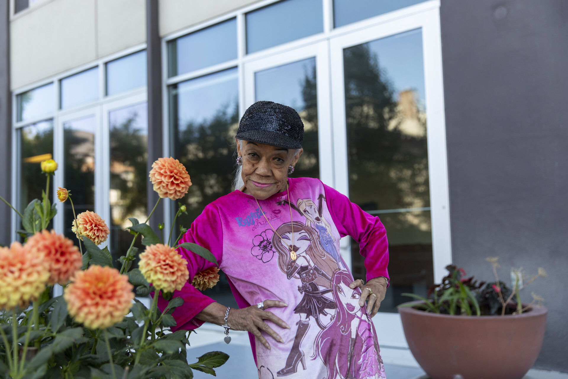 An 87-year-old woman with a pink top and sequined hat poses with her hands on her hips, looking assertively yet playfully at the camera.