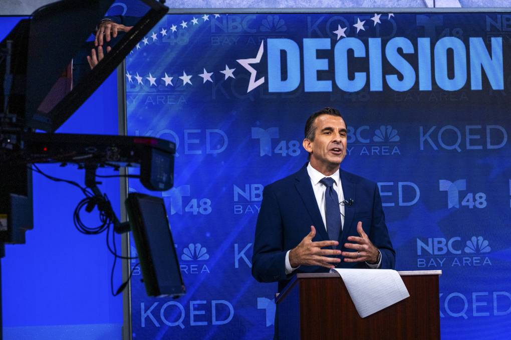 A man in a suit and tie speak behind a podium, with a blue-lit screen behind hime that says 'Decision 2024'