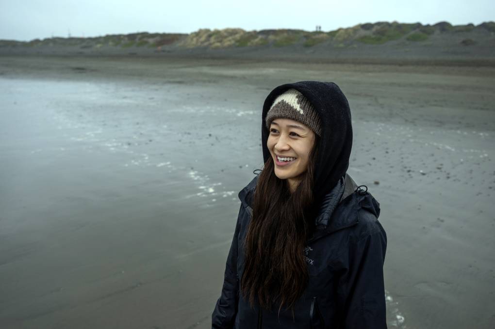 A woman on the beach bundled up in a raincoat and winter beanie smiles as she gazes out toward the sea.