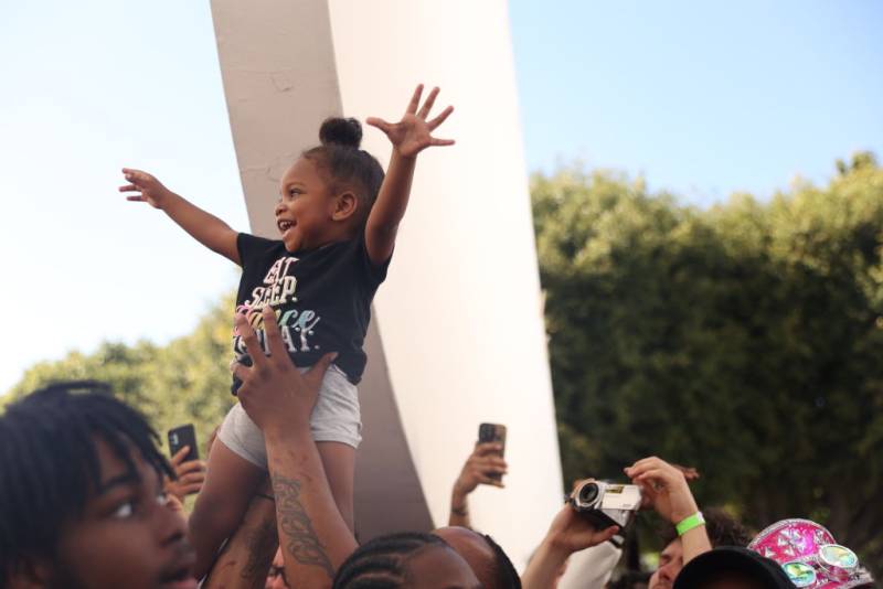 a smiling little girl with brown skin, a black t-shirt and white shorts is hoisted above a crowd during a music video shoot