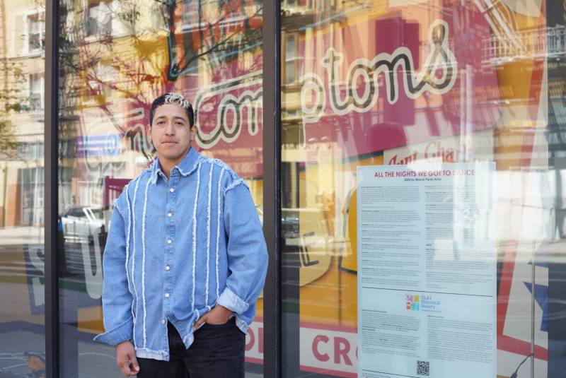 Marcel Pardo Ariza wears a blue button-up shirt while standing in front of their latest work behind a windowfront, "All The Nights We Got To Dance."