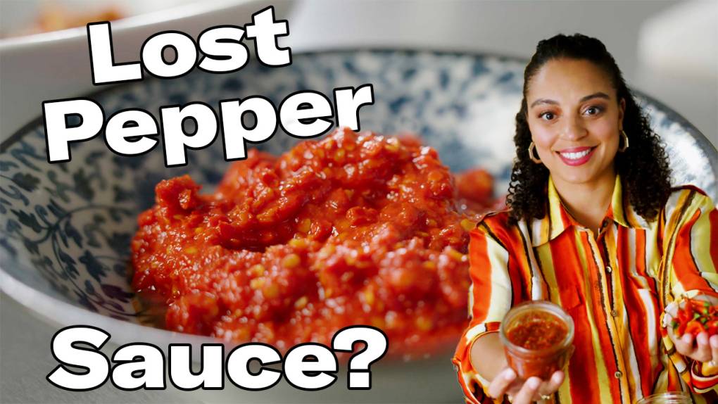 A young light-skinned black woman with long hair holds chile peppers in two outstretched hands. There is a bowl of spicy chile paste behind her.