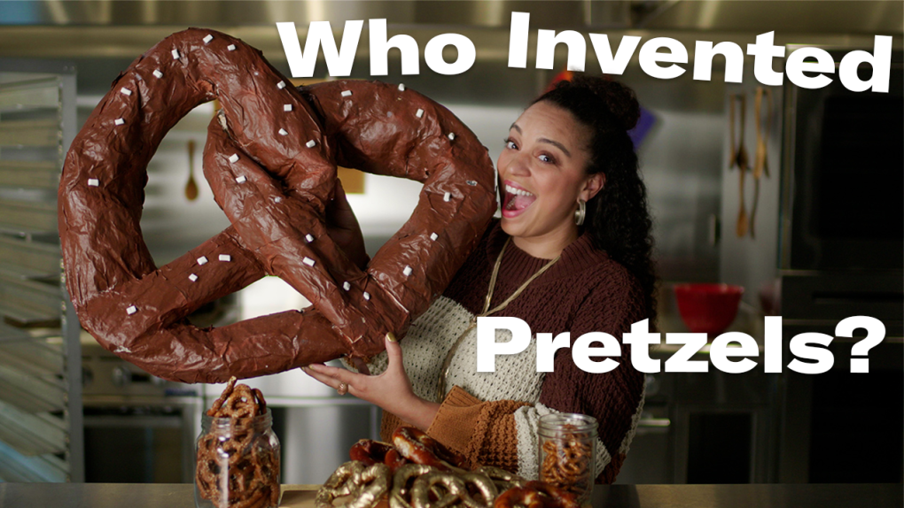 Host of Beyond the Menu, Cecilia Philips is holding a giant paper mache pretzel pretending to eat it. The text on the image reads 'Who Invented Pretzels?'